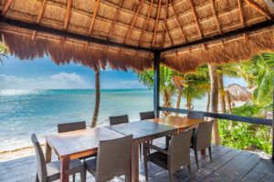 A charming beachfront dining area under a rustic thatched-roof palapa, offering an intimate setting with stunning ocean views. The wooden table and woven chairs invite guests to enjoy a meal while the gentle waves lap against the shore, and the warm sea breeze rustles through the palm trees, creating a serene and romantic atmosphere.