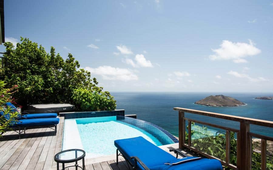 Secluded infinity pool with vibrant blue loungers, overlooking the serene ocean and a small island, surrounded by lush greenery for a private escape.