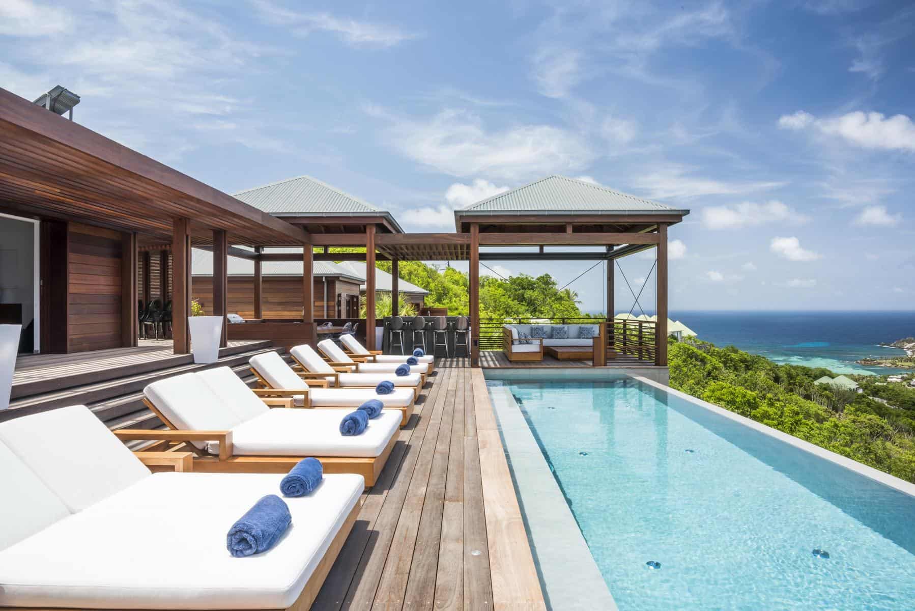 A serene poolside retreat with plush lounge chairs, overlooking vibrant greenery and the sparkling ocean under a bright, cloud-dappled sky.