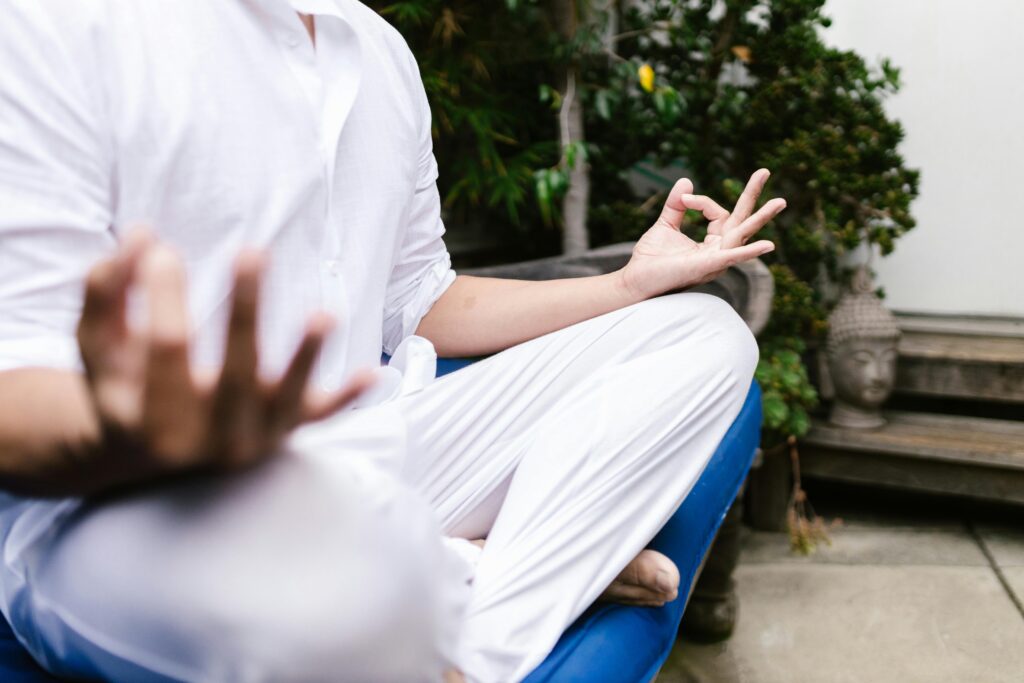 A person dressed in flowing white clothing sits in a peaceful meditation pose, their hands gently forming a mudra. The serene garden setting, with lush greenery and a Buddha statue in the background, enhances the sense of mindfulness and tranquility. Their posture radiates calmness, embodying a moment of deep reflection and inner stillness