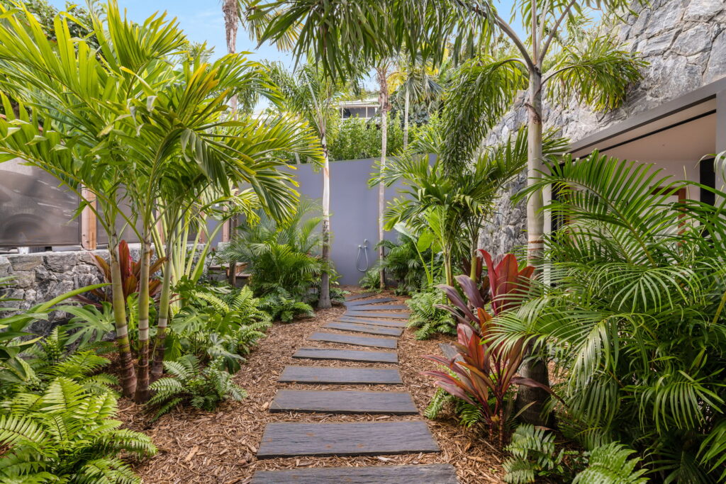 A lush tropical garden pathway in an eco-friendly villa in St. Barts, featuring palm trees, vibrant foliage, and a sustainable outdoor shower. The stone path, surrounded by greenery and natural materials, embodies the island’s commitment to sustainable luxury and harmonious living with nature.