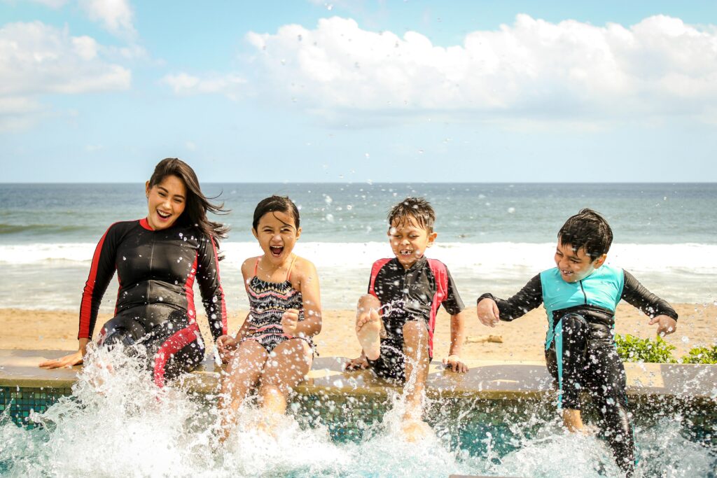 A joyful family moment at the poolside, featuring a woman and three children splashing water with their feet. They are dressed in swimwear and rash guards, smiling and laughing against a scenic beach and ocean backdrop under a bright blue sky