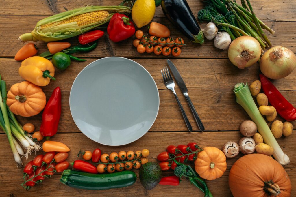 A beautifully arranged assortment of fresh, colorful vegetables and fruits surrounds an empty plate on a rustic wooden table. The display, featuring ingredients like tomatoes, peppers, corn, and pumpkins, reflects the vibrant flavors and farm-to-table philosophy of gourmet kitchens in St. Barts, where local produce is transformed into exquisite culinary creations.