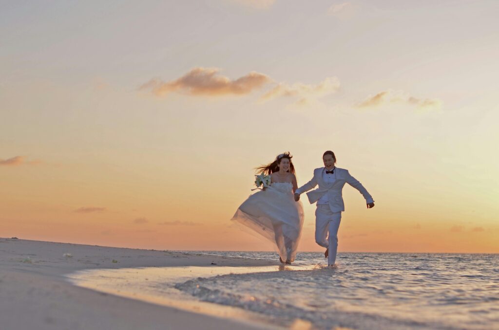 A joyful newlywed couple runs hand in hand along the shoreline of a pristine beach in St. Barts at sunset. The bride’s flowing white gown and the groom’s elegant white suit contrast beautifully against the golden sky, creating a romantic and unforgettable moment. This breathtaking scenery highlights St. Barts as the perfect luxury destination for a dream beach wedding.