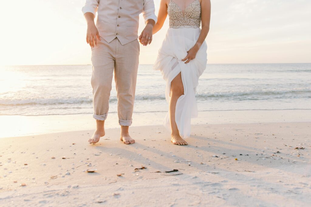 A newlywed couple walks barefoot along a picturesque beach in St. Barts at sunset, holding hands and enjoying a romantic moment. The bride wears a flowing white gown with intricate beadwork, while the groom is dressed in a light linen suit. This serene setting showcases the beauty of St. Barts as a dream wedding destination.