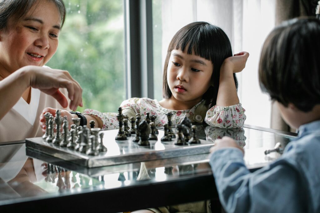 An elderly woman and two children playing chess at a glass table near a window. The young girl in a floral dress looks deep in thought, resting her head on her hand, while the boy in a blue shirt watches attentively. The woman is smiling as she makes a move on the metallic chessboard.